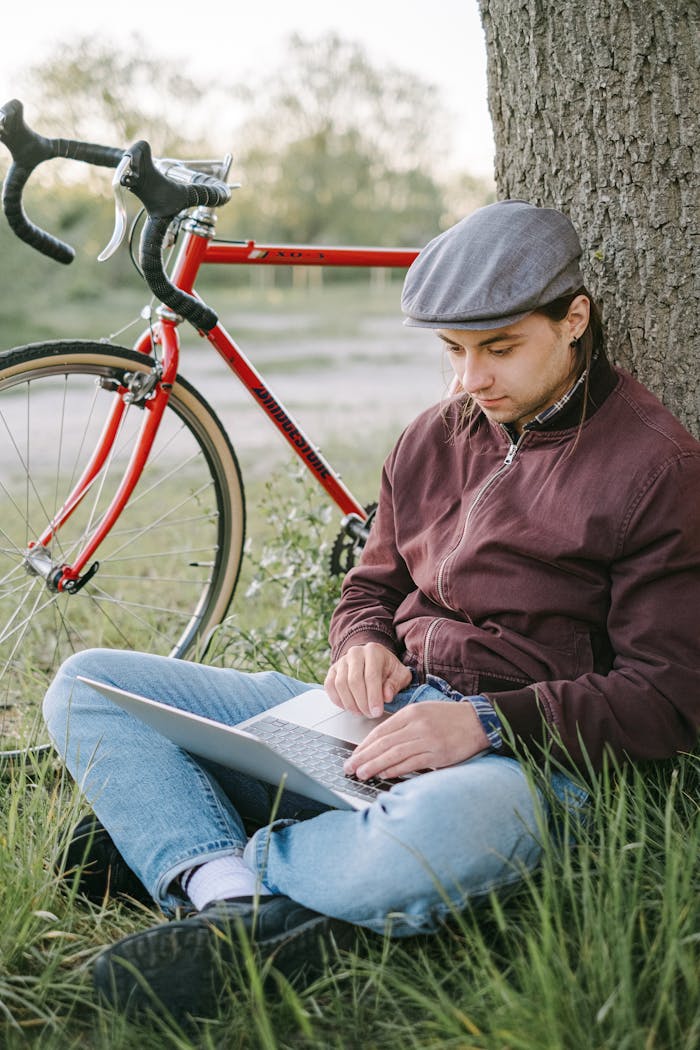 Young man sitting under a tree working on a laptop, next to a bicycle in a grassy outdoor setting.